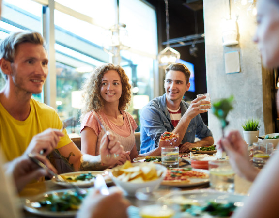 Cheerful young friendly people having talk by table served with tasty meal in cafe or restaurant
